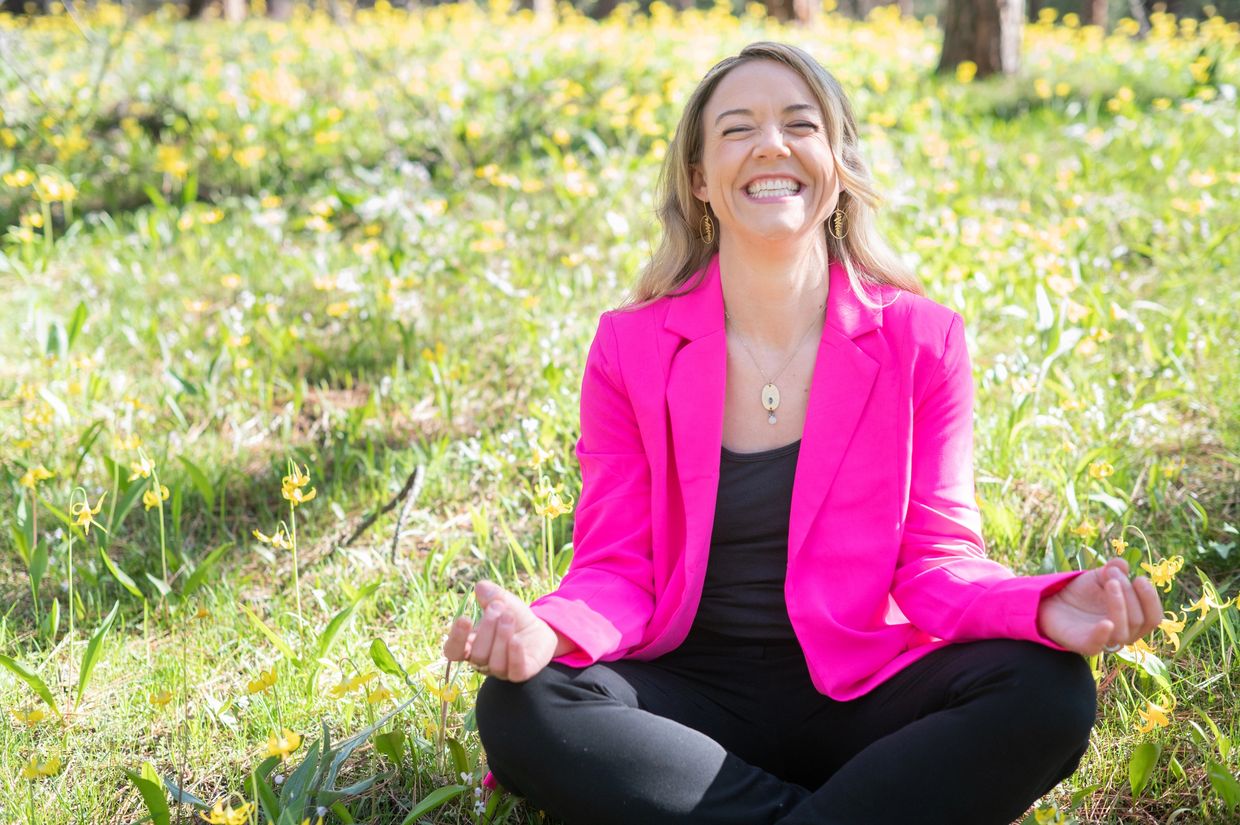 A woman in a bright pink blazer meditates joyfully in a field of yellow flowers.