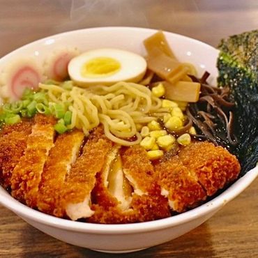A steaming bowl of ramen topped with crispy chicken, half a boiled egg, corn, seaweed, and green onions.