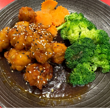 Sesame-coated crispy chicken with broccoli and carrot slices on a dark plate.