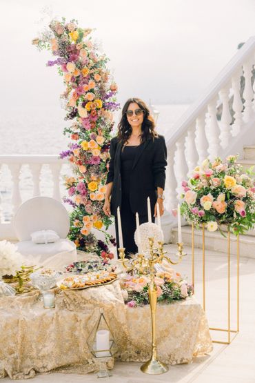 A woman in black stands Beside a floral arch and elegant table setup with candles by the seaside.