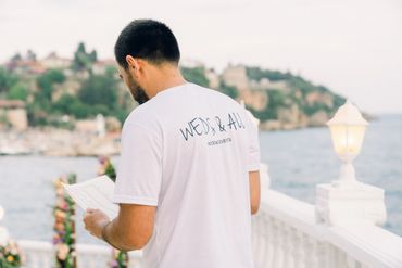 Man reading notes by the seaside wearing a 'WEDS & ALL' event shirt. Antalya Wedding Planner