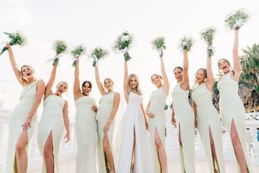 Bride and bridesmaids in elegant white dresses holding bouquets, celebrating joyfully outdoors.