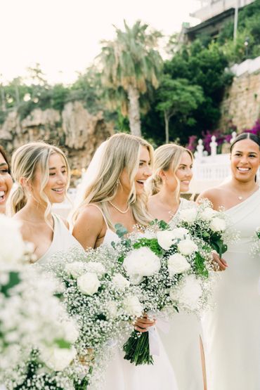 Bridesmaids in white dresses holding lush white floral bouquets outdoors.