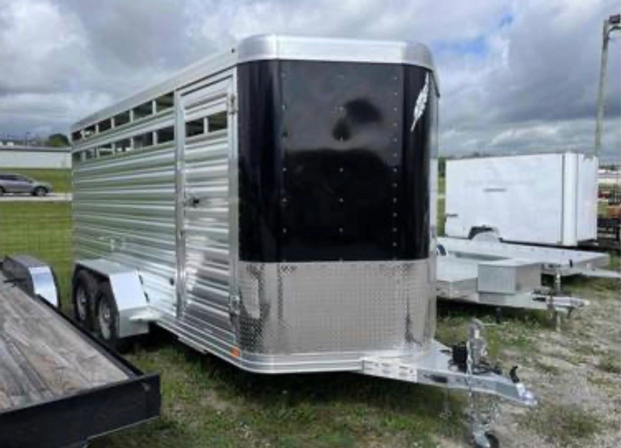 A silver and black livestock trailer parked on grass with other trailers nearby.