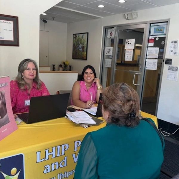 Two women assist a visitor at an informational booth with a yellow tablecloth.