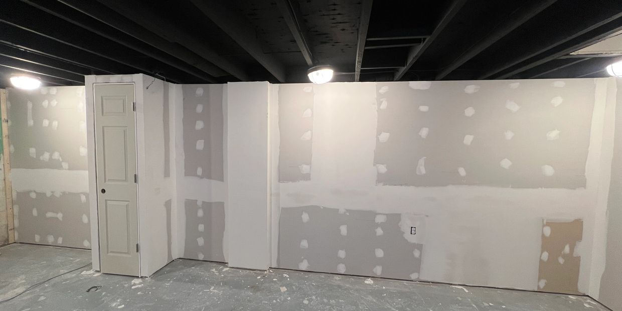 Basement room with unfinished drywall and exposed ceiling beams.