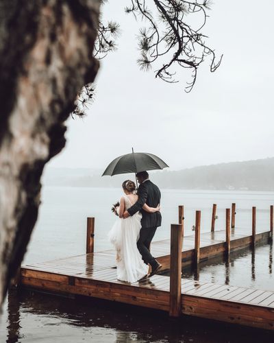 Wedding couple walking in the rain holding an umbrella on the dock near a lake