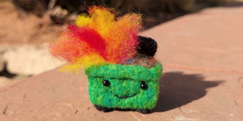 A cute felted green dumpster with a smiling face and colorful flames.