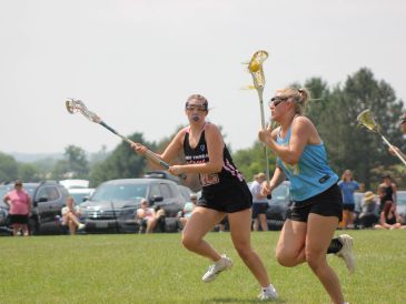 Two female lacrosse players compete on a sunny field.