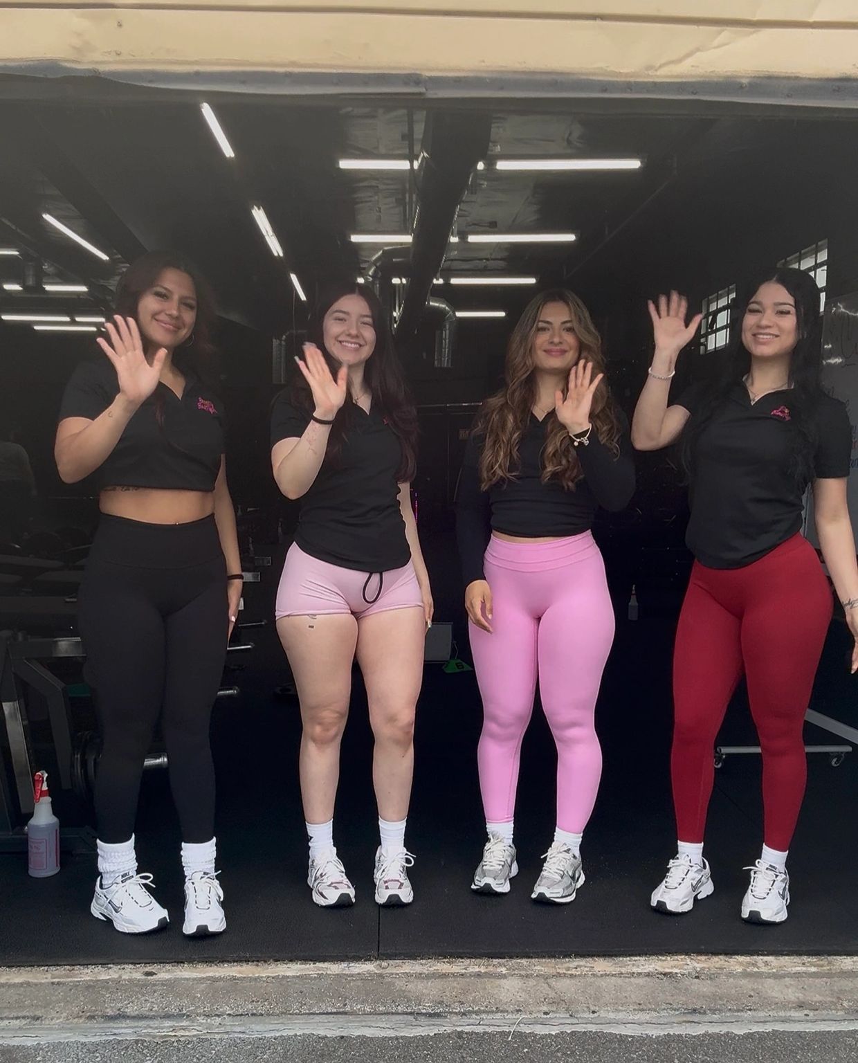 Four women in workout attire waving and smiling in a gym entrance.