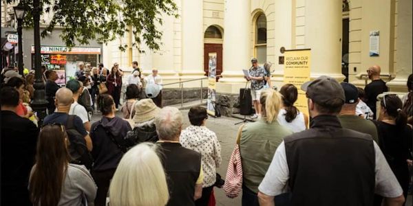Crowd gathered outdoors listening to a speaker near a historic building.