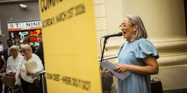 Woman speaking into microphone at outdoor event with audience.