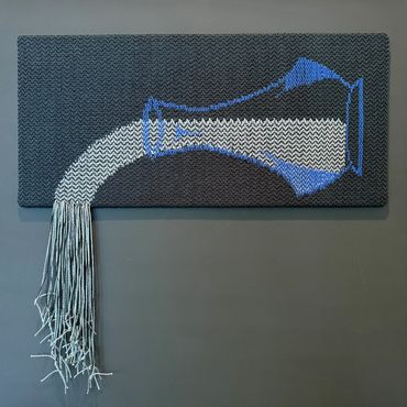 Textile art of a tilted jug pouring out threads onto a wall.