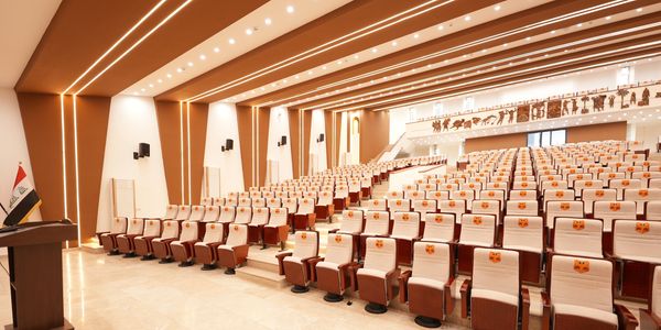 A modern, spacious auditorium with beige and brown seating and an Iraqi flag on the podium.