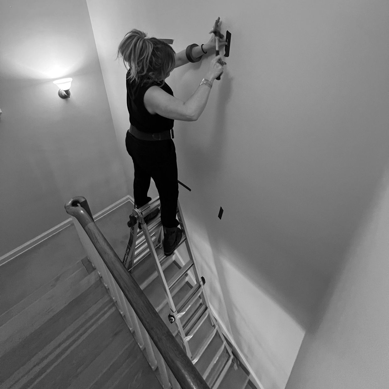 Person standing on a ladder, hammering a nail into a wall in a stairwell.