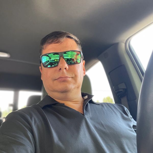 Man wearing reflective sunglasses sitting in a car, taking a selfie.