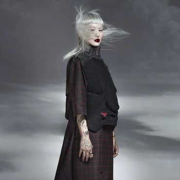 Person with white hair and tattoos wearing dark clothes in gray fog.