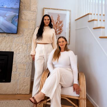 Two women in neutral-toned outfits posing in a cozy, modern living room with natural decor.