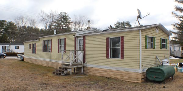 A beige mobile home with maroon shutters and a small porch in a rural area.