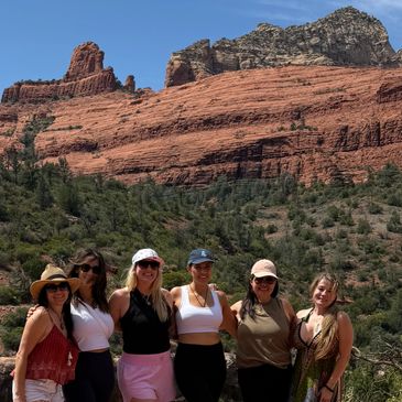 Group of six women posing in front of majestic red rock formations and green vegetation.