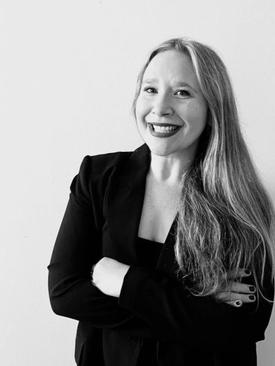 Smiling woman with long hair in a black blazer poses confidently against a plain background.