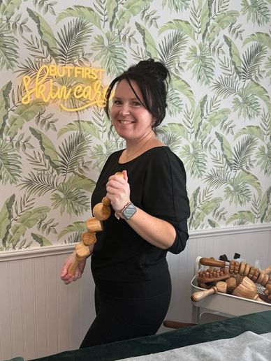 Smiling woman holding wooden massage tools in a spa room with tropical wallpaper.
