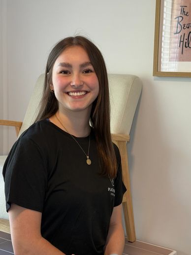 Smiling young woman sitting indoors with a framed Beverly Hills poster behind her.