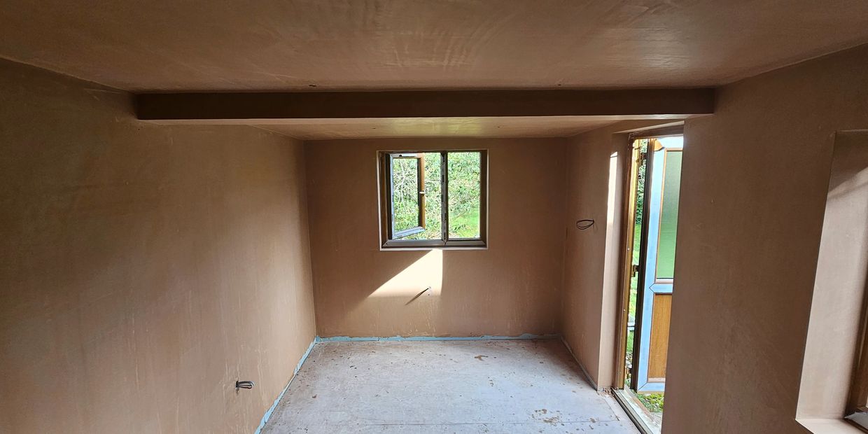 Empty room with fresh plaster walls, an open window, and a door leading outside.