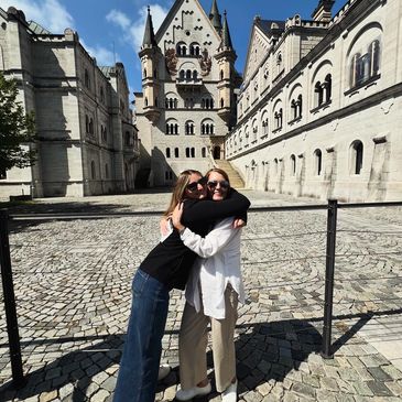 Two women hugging in front of a historic castle on a sunny day.