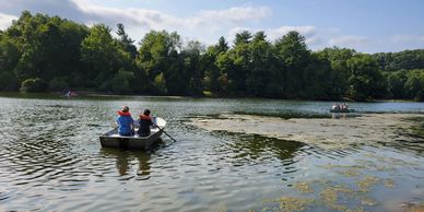Two people rowing a boat on a calm lake surrounded by trees.
