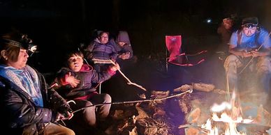 People roasting food and relaxing around a campfire at night.