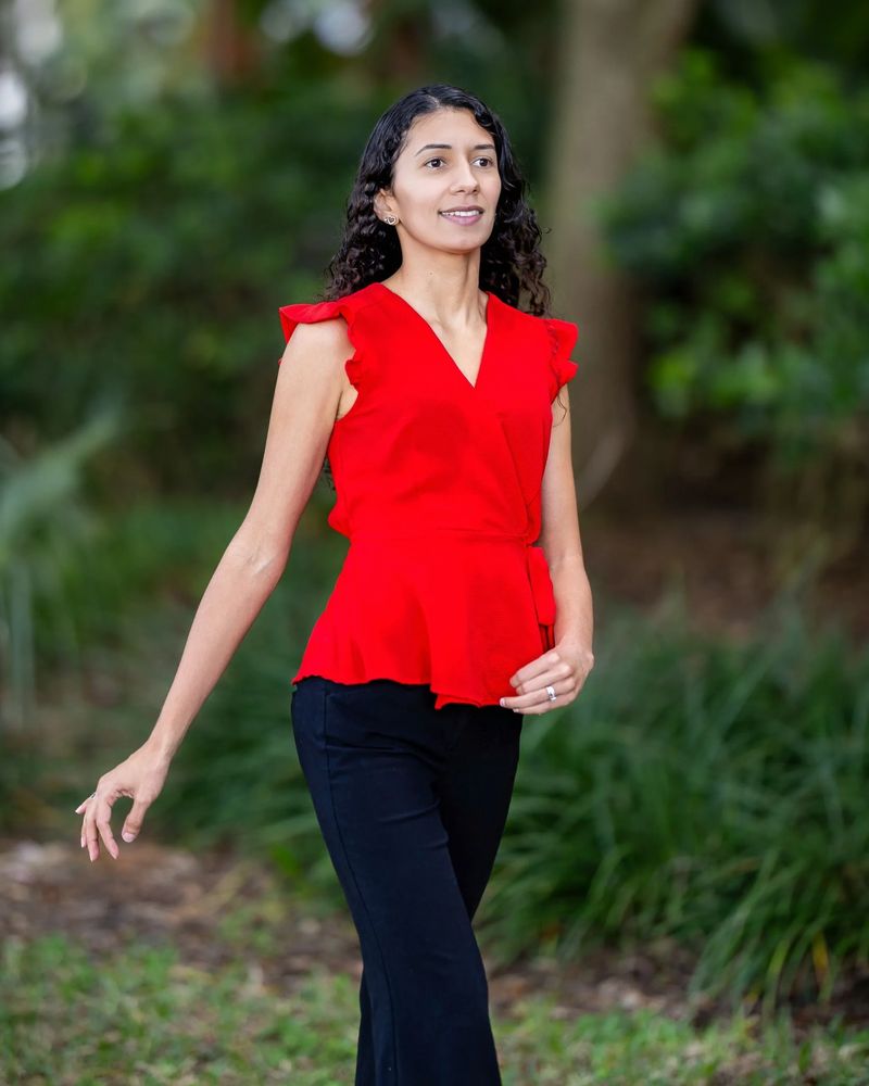 Woman in a red sleeveless blouse and black pants walking outdoors.