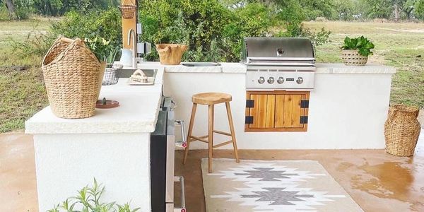 Cozy outdoor kitchen with grill, wooden stool, and greenery under a wooden pergola.