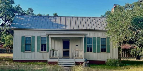 Small white house with green shutters and metal roof under clear blue sky.