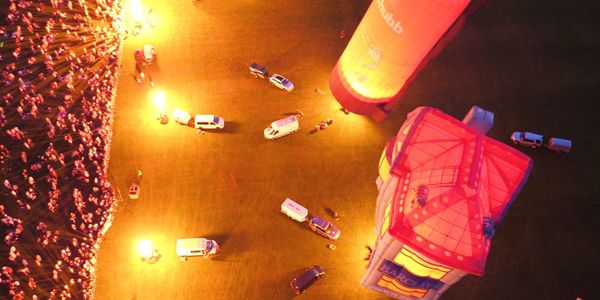 Aerial view of a crowd and vehicles illuminated by bright lights near a large inflatable structure at night.