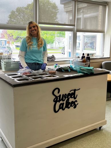 Woman serving desserts at a Sweet Cakes counter in a bright room.