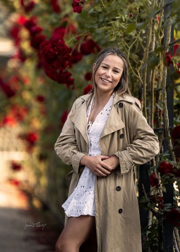 Junge Frau mit beige Trenchcoat vor roten Blumen stehend.