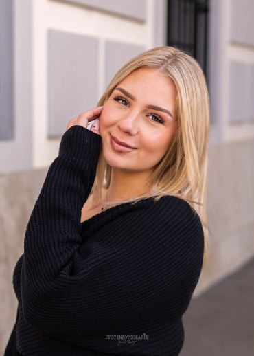 Junge blonde Frau mit schwarzem Pullover lächelt.