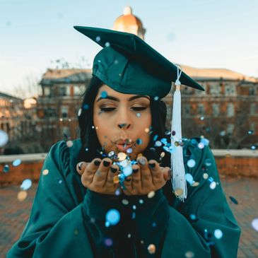 Graduate in cap and gown blowing blue confetti outdoors.