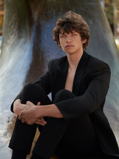 Young man in a black suit sitting with arms around knees outdoors.