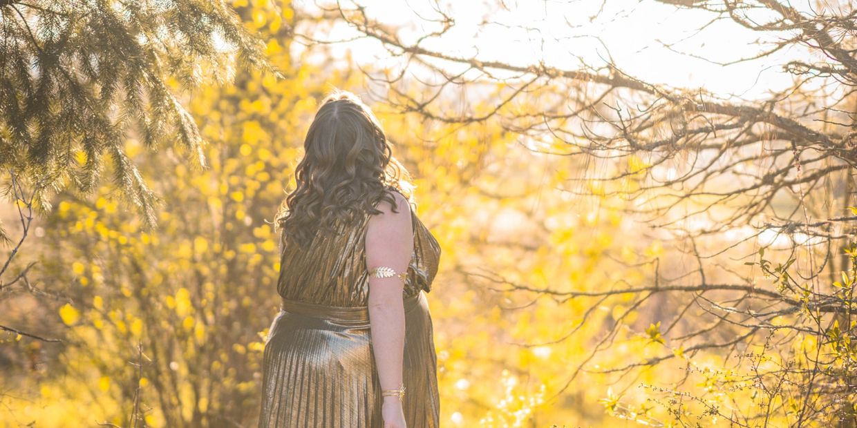 Woman in a golden dress stands amidst sunlit autumn trees.