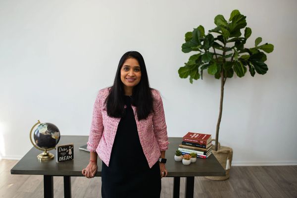 A woman stands confidently by a desk with books, a globe, and a plant in a bright, minimalist room.