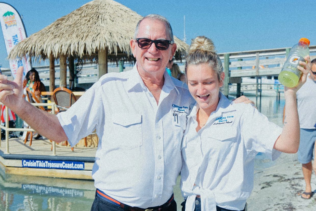 Two cheerful people in white shirts pose by a tiki hut near the water.