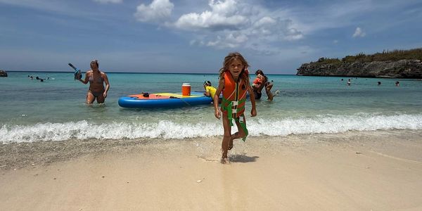 A child in a life jacket runs on a sandy beach with people enjoying the ocean behind.
