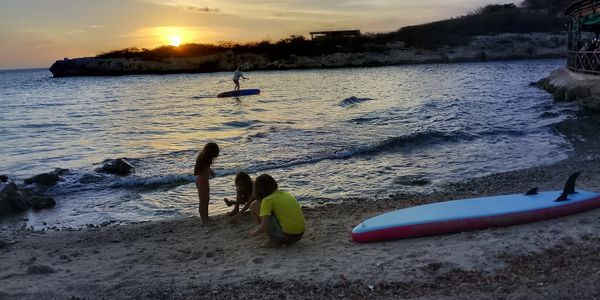 Children playing on the beach at sunset with a paddleboarder in the water.