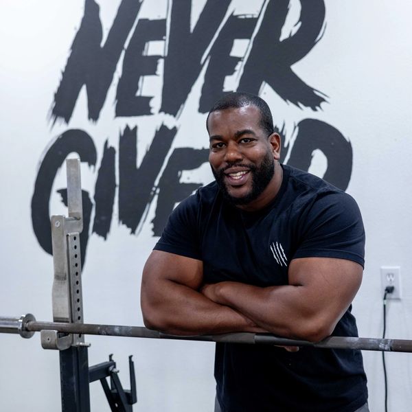 Smiling man in gym with 'Never Give Up' mural behind him.