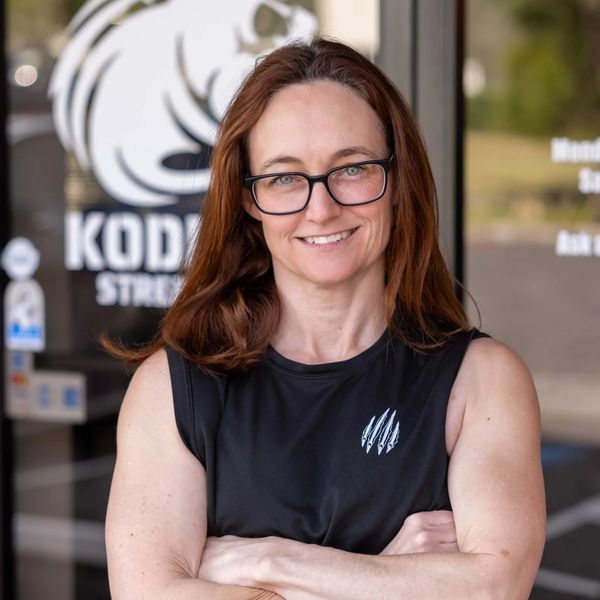 A confident woman with glasses and brown hair stands with arms crossed in front of a fitness center.