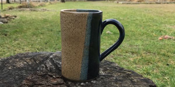 A ceramic mug with a speckled and blue glaze on a tree stump outdoors.