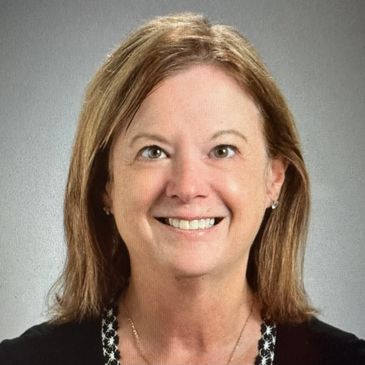 Smiling woman with light brown hair and a black top against a gray background.