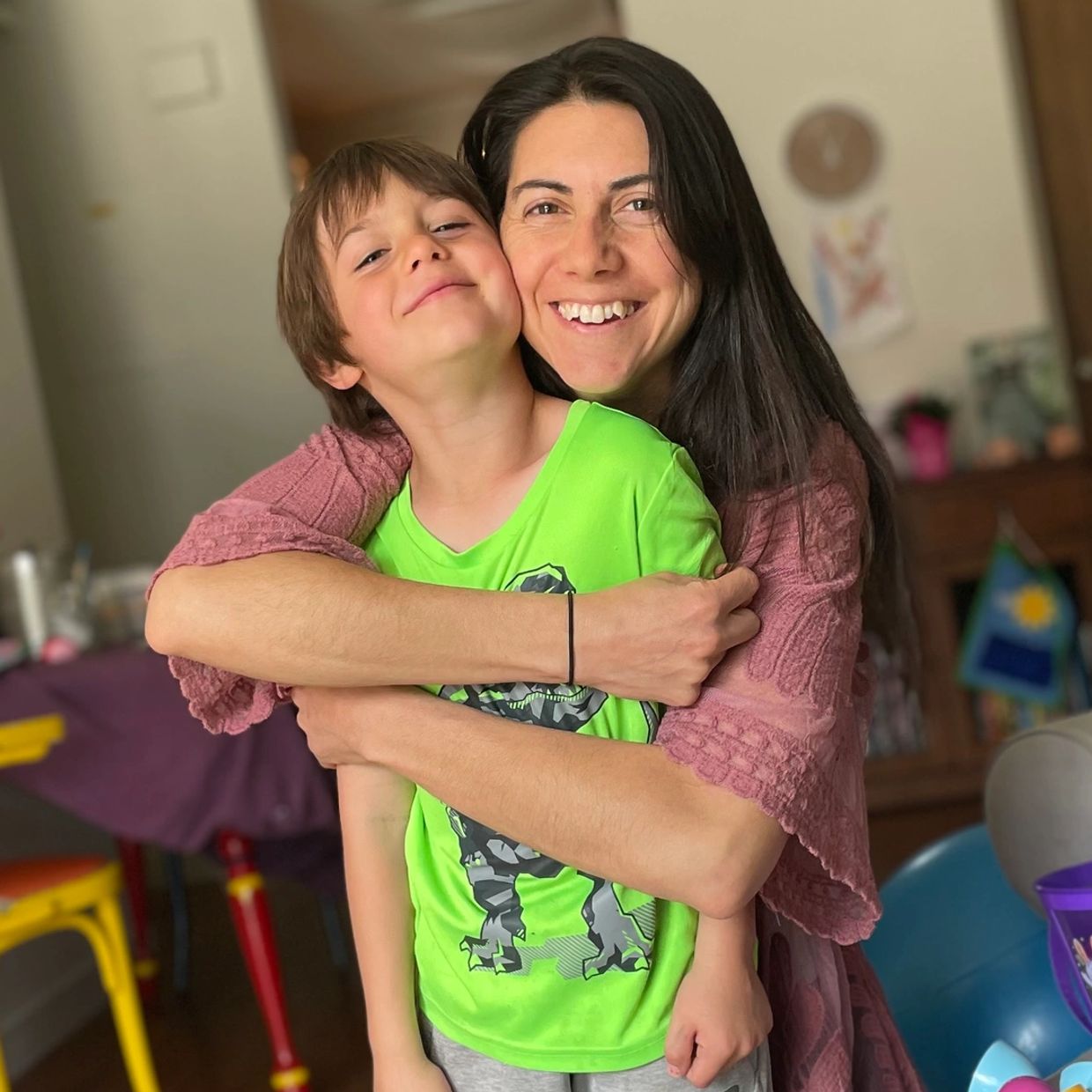 A woman happily hugs a smiling boy wearing a green dinosaur t-shirt.